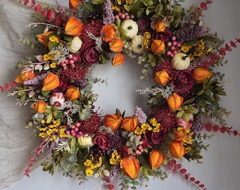 May include: A circular wreath featuring a dense arrangement of artificial orange Chinese lanterns, small white pumpkins, deep burgundy roses, yellow wildflowers, pink berries, and green eucalyptus foliage with long, thin red-leafed stems.