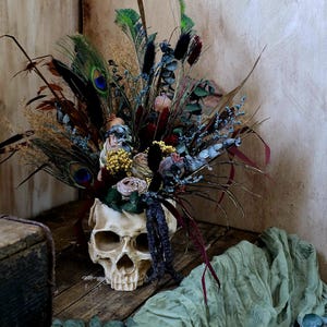 May include: A human skull with a dried flower arrangement inside. The skull is white and has a detailed bone structure. The flowers are a mix of dried grasses, feathers, and small pink and white flowers. The skull is sitting on a wooden surface with a green cloth and eucalyptus leaves in the foreground.