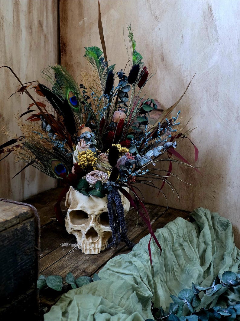 May include: A human skull with a dried flower arrangement inside. The skull is white and has a detailed bone structure. The flowers are a mix of dried grasses, feathers, and small pink and white flowers. The skull is sitting on a wooden surface with a green cloth and eucalyptus leaves in the foreground.