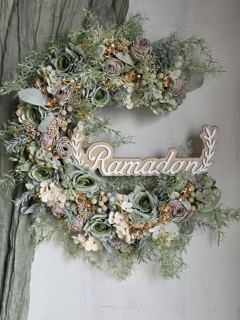 May include: A crescent-shaped floral wreath with the word "Ramadan" in white script. The wreath features green, cream, and mauve flowers, gold accents, and greenery. The background is a light-coloured wall with a green fabric.