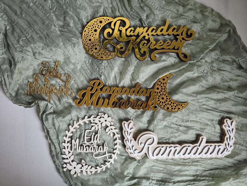May include: Gold and white Ramadan and Eid Mubarak decorations. The gold pieces include the phrases "Ramadan Kareem" and "Ramadan Mubarak" with a crescent moon design. The white pieces include the phrases "Eid Mubarak" and "Ramadan" with floral and leaf accents.