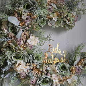 May include: A crescent-shaped floral wreath with green, cream, and mauve roses, complemented by gold-coloured foliage and berries. A gold script reads "Eid Mubarak". This decorative piece is suitable for celebrations.