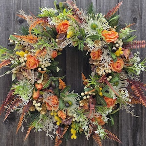May include: A circular wreath with orange roses, white berries, and diverse green and brown foliage. The wreath is decorated with ferns and other elements, creating a natural, autumnal look. It is displayed against a weathered wooden background, with a rustic feel.