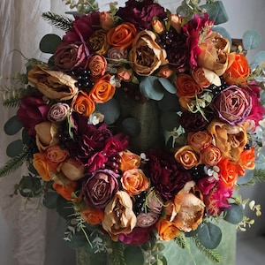 Peut inclure: Une couronne florale vibrante composée de fleurs orange, bordeaux et beige, agrémentée de feuillage vert et de baies foncées. La couronne circulaire est parfaite pour la décoration saisonnière.