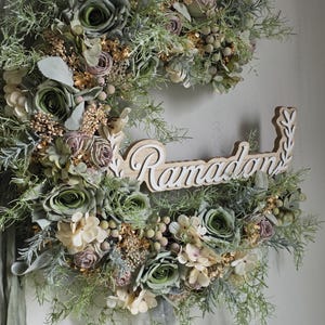 May include: A crescent-shaped floral wreath featuring green, cream, and mauve flowers, with gold and green foliage. The wooden sign in the centre reads "Ramadan" in a decorative font, suitable for seasonal home decor.