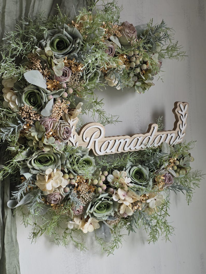 May include: A crescent-shaped floral wreath featuring green, cream, and mauve flowers, with gold and green foliage. The wooden sign in the centre reads "Ramadan" in a decorative font, suitable for seasonal home decor.