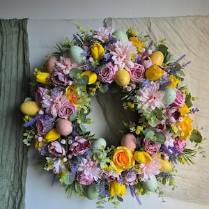 May include: A vibrant Easter wreath featuring a circular arrangement of colorful flowers, including pink dahlias, yellow tulips, and purple roses. Pastel-colored speckled eggs are interspersed throughout the floral design, creating a festive spring decoration.
