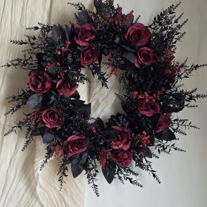May include: A circular wreath featuring deep burgundy and black roses interspersed with dark black foliage, thin sprigs, and small dark berries. The arrangement is densely packed with dark leaves and floral elements, creating a full, rounded shape with a consistent colour palette of black and dark red throughout the entire decorative piece.