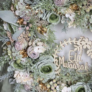 May include: A decorative wreath with faux flowers in green, cream, and pale pink. The wreath includes sprigs of greenery and gold accents. The words "Eid Mubarak" are cut from wood and integrated into the design, celebrating Eid.