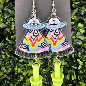 May include: A pair of novelty earrings featuring a UFO design with a rainbow pattern and the text "NO BAD TRIPS". The earrings have a silver hook and a neon green earplug charm.
