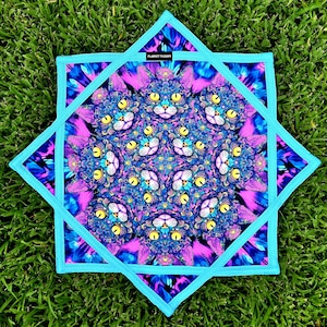 May include: A vibrant, star-shaped quilted pot holder featuring a repeating pattern of cat faces in shades of blue, purple, and yellow. The design is framed by a light blue border and a darker blue edge. The text "PLANET STUDIOS" is visible.