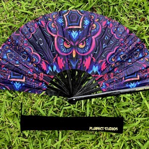 May include: A decorative folding fan with a vibrant owl design in shades of purple, pink, and blue. The fan is open, revealing the intricate artwork. The fan's black ribs are visible. A black band with gold text that reads "PLURFECT STUDIOS" is at the bottom.