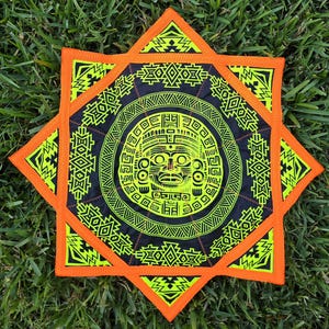 May include: A decorative, star-shaped textile with a central circular design featuring a stylized face in neon green against a black background. The star has orange edges and neon green geometric patterns. The textile is placed on a bed of green grass.