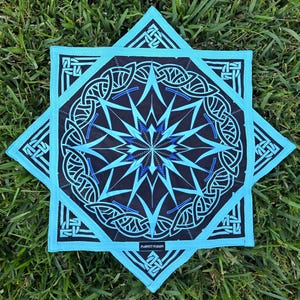 May include: A square, black and turquoise bandana with a geometric design. The center features a starburst pattern with blue accents. The border has a turquoise frame with a Celtic knot design. The text "PLURFECT STUDIOS" is printed on the bottom.