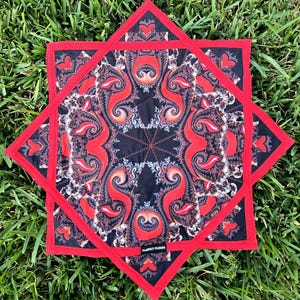 May include: A decorative, star-shaped textile with a red border and a black and red paisley pattern. The center features a complex, symmetrical design. The text "PLANET GLOW" is visible on the bottom edge. The textile is displayed on green grass.