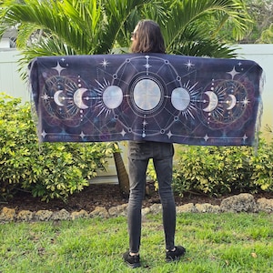 May include: A person wearing a black tapestry with a moon phases design and geometric patterns. The tapestry is draped over their shoulders and has a fringe border. The tapestry is printed with the text "PURPLE STUDIO".