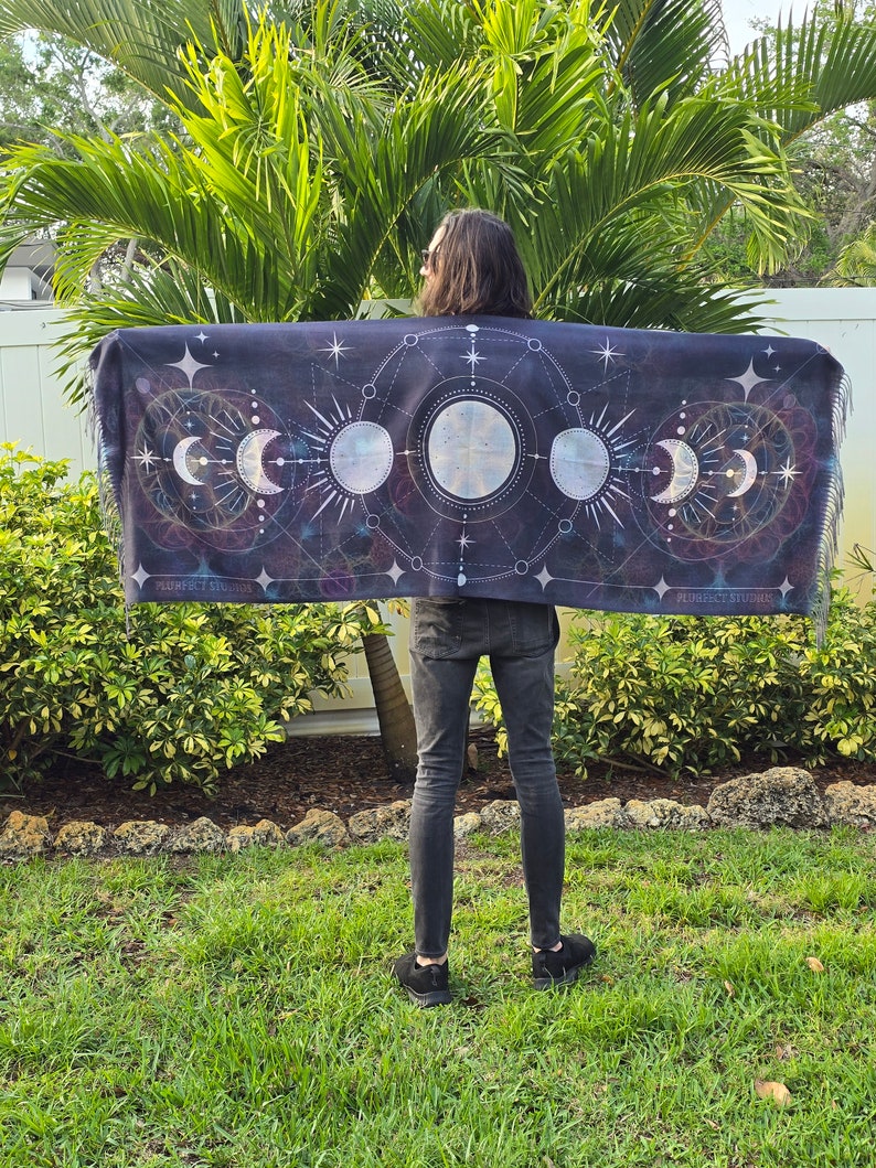 May include: A person wearing a black tapestry with a moon phases design and geometric patterns. The tapestry is draped over their shoulders and has a fringe border. The tapestry is printed with the text "PURPLE STUDIO".
