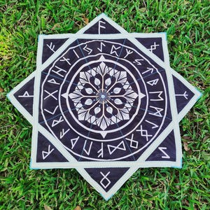 May include: A black and white decorative square with a star-like shape. The design features a central floral pattern surrounded by a ring of runes. The edges are outlined in a light blue color, and the background is green grass.