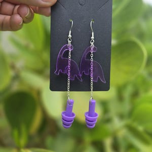 May include: Purple dinosaur-shaped earrings with silver chain accents and purple earplugs. The earrings are displayed on a black card, held against a blurred green background.