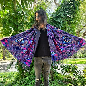 May include: A person wearing a black shirt and blue jeans is standing in front of a green bush. They are wearing a large, colourful shawl with a fringe border. The shawl has a psychedelic pattern with geometric shapes and bright colours.