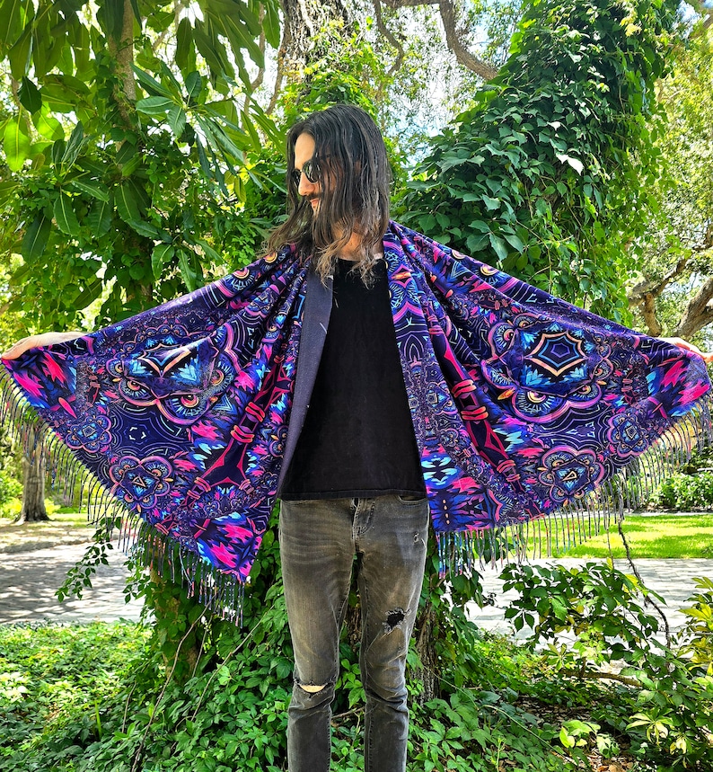 May include: A person wearing a black shirt and blue jeans is standing in front of a green bush. They are wearing a large, colourful shawl with a fringe border. The shawl has a psychedelic pattern with geometric shapes and bright colours.