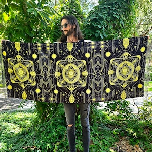 May include: A black and gold tapestry with geometric patterns and a central design of a star within a circle. The tapestry is held up by a person in front of a green leafy background.
