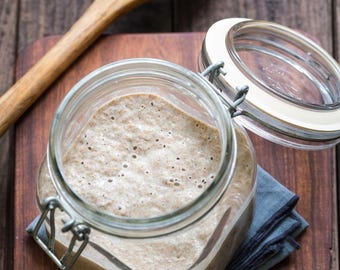 May include: A glass jar with a lid, filled with a light brown, bubbly mixture. The jar is sitting on a wooden surface.