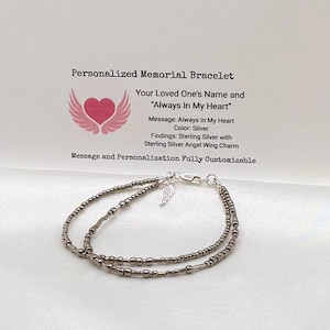 Op de afbeelding: Een zilverkleurige kralenarmband met een kleine zilveren engelenvleugelbedel en een karabijnsluiting. De armband wordt getoond voor een kaart met de tekst Personalized Memorial Bracelet, Your Loved One's Name and Always In My Heart.