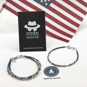 May include: Two bracelets made of small dark blue and metallic silver beads with silver-toned clasps. A black card displays the white logo of a hat and the text code secret Custom Morse Code Jewelry. A circular sticker features the Space Force logo.