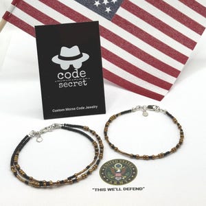 May include: Two bracelets made of small black and bronze beads with silver-toned clasps and heart charms. A black display card features a white hat icon and the text code secret Custom Morse Code Jewelry. A United States Army logo is visible.