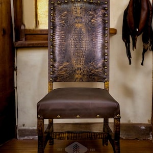 May include: A brown leather chair with a crocodile pattern on the back and seat. The chair has a wooden frame and brass studs. The chair is sitting on a wooden floor. The text "EST. 1989 RUSTIC EXPRESSIONS&reg;" is visible on the floor.