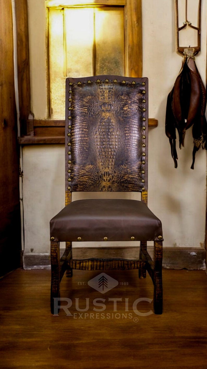 May include: A brown leather chair with a crocodile pattern on the back and seat. The chair has a wooden frame and brass studs. The chair is sitting on a wooden floor. The text "EST. 1989 RUSTIC EXPRESSIONS&reg;" is visible on the floor.