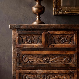 May include: A rustic wooden dresser with intricate carved floral designs on the drawers. A bronze-colored lamp with a beige lampshade sits on top. A gold-framed mirror hangs in the background, adding a touch of elegance.