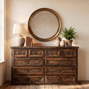 May include: A dark brown wooden dresser with carved floral designs on the drawers. A round gold mirror hangs above the dresser, which is topped with books, pottery, a lamp, and a potted fern. The dresser sits on a wood floor with a woven rug.