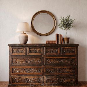 May include: A dark brown wooden dresser with carved floral and scroll designs. A round gold mirror hangs above the dresser, with a lamp, books, and a potted plant on top. The word "RUSTIC" is visible on the bottom of the dresser.