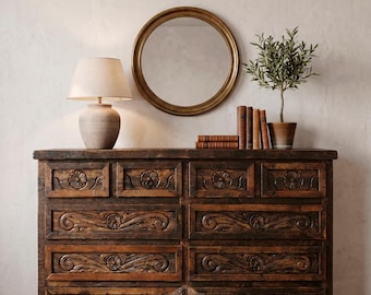 Può includere: Una cassettiera in legno marrone scuro con intagli floreali e a volute. Uno specchio rotondo dorato è appeso sopra la cassettiera, con una lampada, libri e una pianta in vaso. La parola "RUSTIC" è visibile in basso.