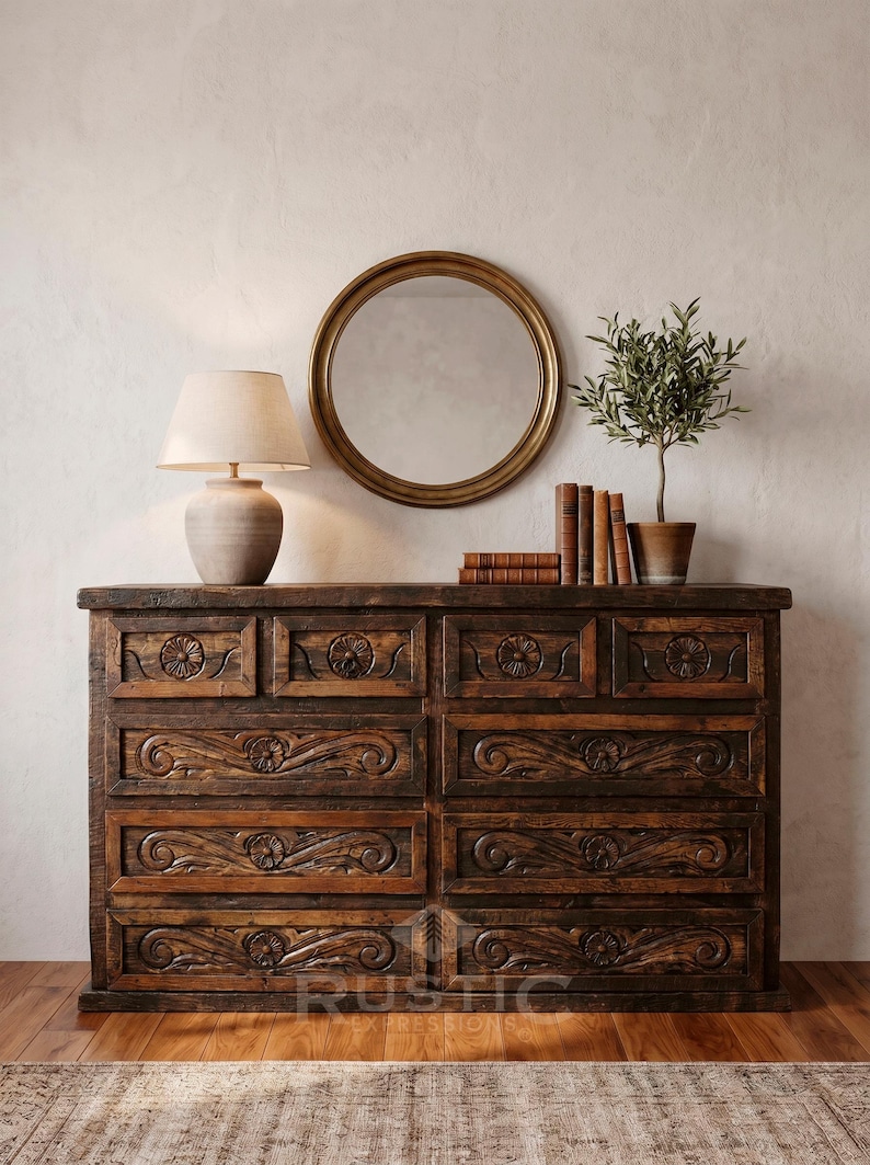 May include: A dark brown wooden dresser with carved floral and scroll designs. A round gold mirror hangs above the dresser, with a lamp, books, and a potted plant on top. The word "RUSTIC" is visible on the bottom of the dresser.