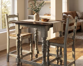May include: A high-top dining set featuring a square table with a dark brown wooden top and distressed light gray legs, accompanied by two matching ladder-back chairs with woven seats, a beige table runner, a ceramic vase, and a wooden bowl.
