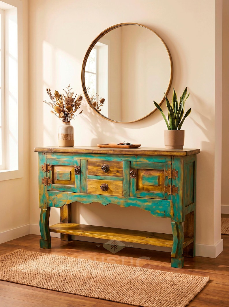 May include: A rustic wooden console table with turquoise and yellow paint, featuring drawers and cabinet doors. A round gold mirror hangs above, with a vase of dried flowers and a potted plant on the table.