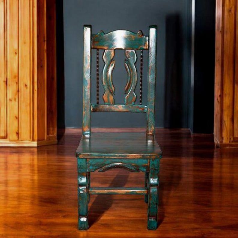 Puede incluir: Una silla de madera azul verdosa con respaldo tallado y acabado desgastado. La silla tiene un dise&ntilde;o sencillo con un asiento rectangular y cuatro patas.