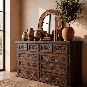 May include: A dark brown wooden dresser with carved floral designs on the drawers. The dresser is topped with a round mirror, pottery, and a potted plant. The room has a rustic, warm aesthetic.