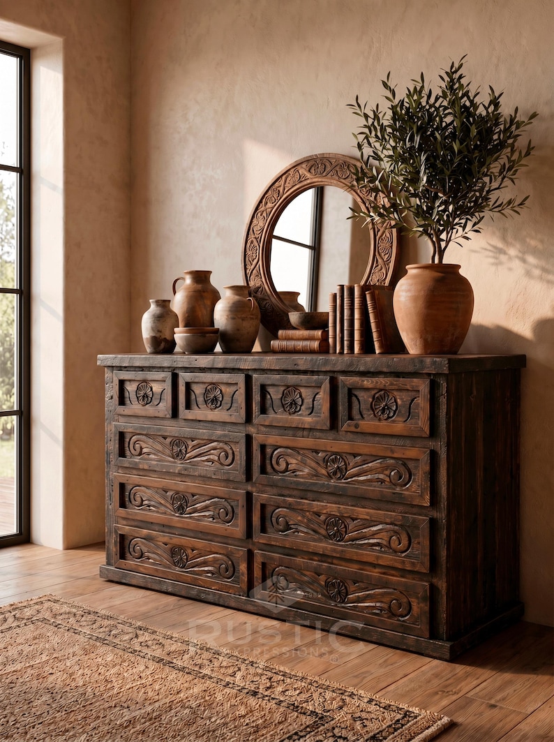 May include: A dark brown wooden dresser with carved floral designs on the drawers. The dresser is topped with a round mirror, pottery, and a potted plant. The room has a rustic, warm aesthetic.