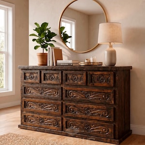 May include: A dark brown wooden dresser with carved floral and scroll designs on the drawer fronts. A round gold mirror hangs above the dresser, reflecting a window. A lamp with a beige shade and a potted plant sit on top.