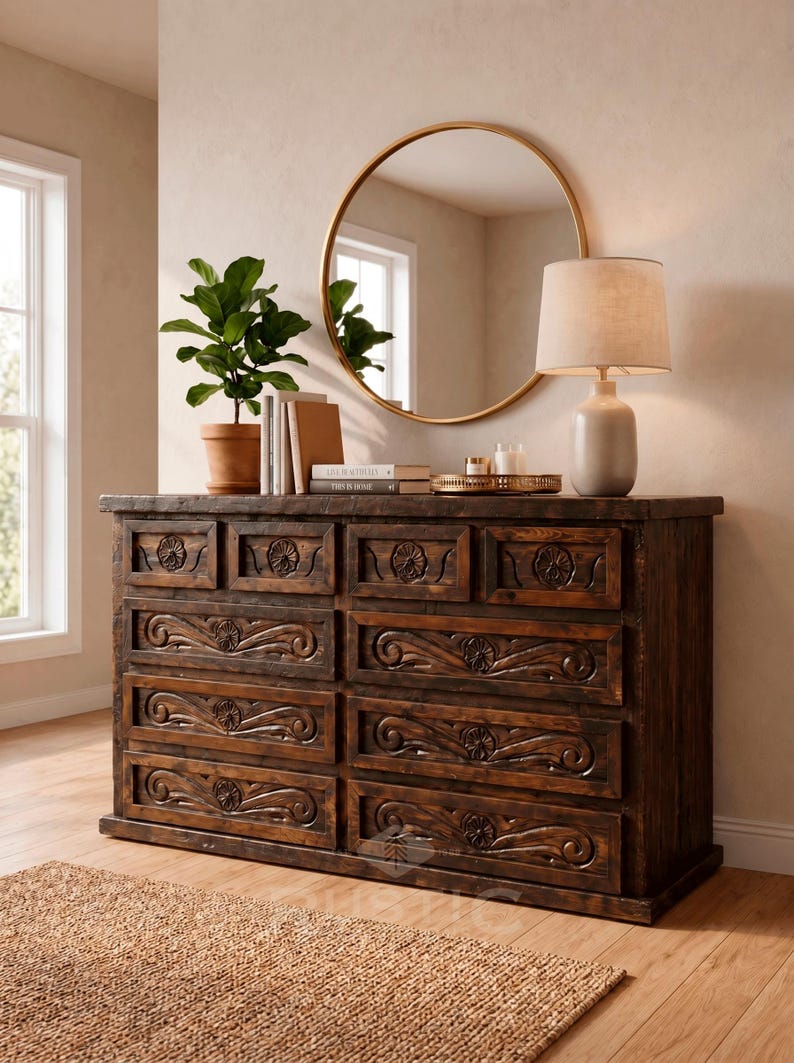 May include: A dark brown wooden dresser with carved floral and scroll designs on the drawer fronts. A round gold mirror hangs above the dresser, reflecting a window. A lamp with a beige shade and a potted plant sit on top.