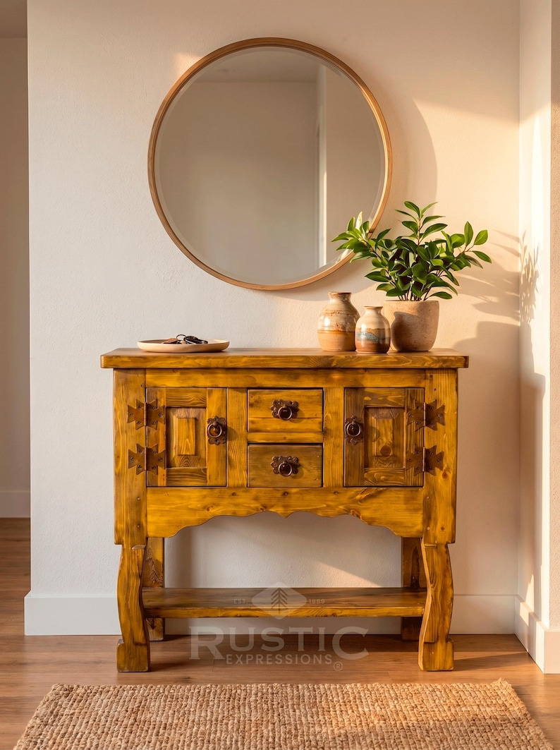 May include: A rustic wooden console table with two drawers and two cabinet doors, adorned with decorative hardware. A round mirror hangs above the table, and a small plant and vases sit on top. The table is made of light brown wood and has a lower shelf.
