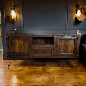 May include: A rustic, wooden media console with a distressed finish. The cabinet has two doors, a drawer, and an open shelf. It stands on black hairpin legs. The console is set against a dark gray wall with sconces.