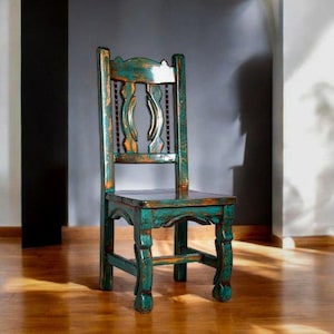 Puede incluir: Una silla de madera azul verdosa con un respaldo tallado y pintura desgastada. La silla tiene un dise&ntilde;o sencillo con un asiento rectangular y cuatro patas.