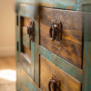 May include: A rustic wooden cabinet with two drawers and a side panel. The cabinet features a distressed finish with shades of brown, yellow, and teal. The drawers have ornate metal ring pulls.