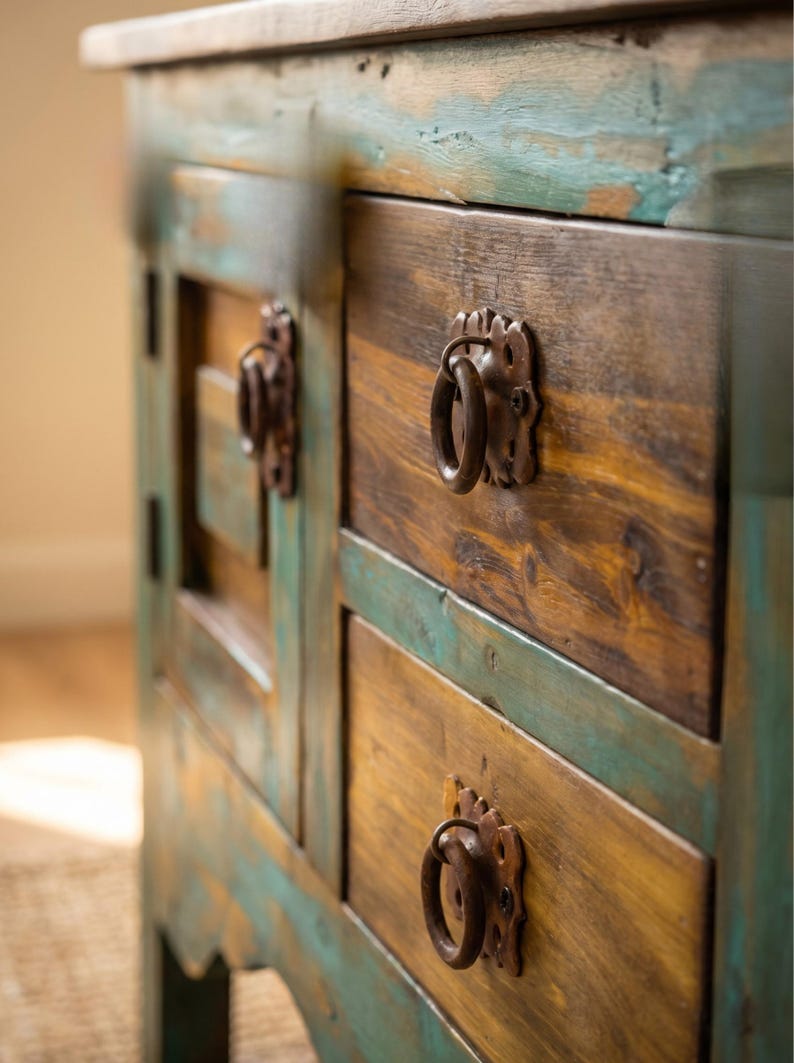 May include: A rustic wooden cabinet with two drawers and a side panel. The cabinet features a distressed finish with shades of brown, yellow, and teal. The drawers have ornate metal ring pulls.