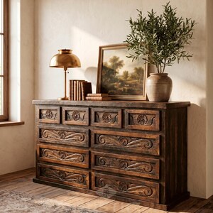May include: A dark brown wooden dresser with carved floral and scroll details on the drawers. A brass lamp, books, a framed landscape painting, and a vase with greenery sit on top. The dresser is in a room with a wooden floor.